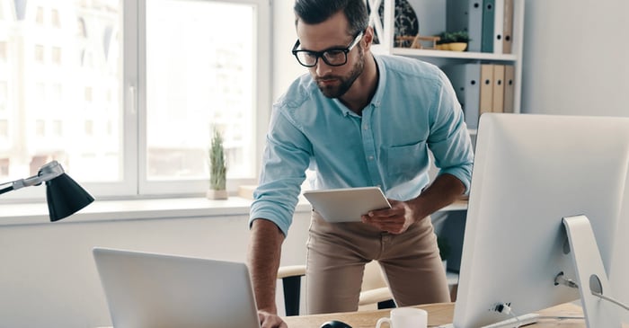 A man wearing glasses, a blue shirt, and glasses, is holding an iPad and looking at a laptop